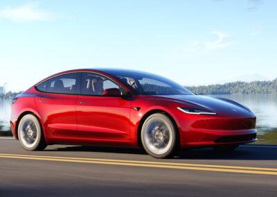 czerwona tesla 3 jedzie drogą obok jeziora, widać prawy profil i przód auta.