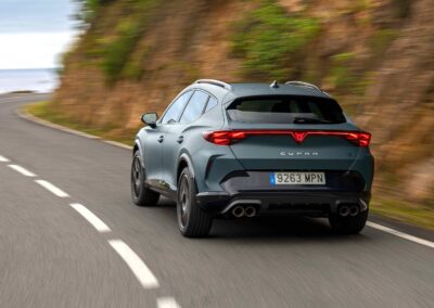 Niebieska cupra formentor jadąca drogą asfaltową na zakręcie, z boku skalne urwisko, w oddali ocean. Widać tył i lewy bok samochodu.