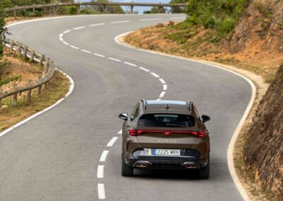 Tył brązowej cupry formentor jadącej drogą wśród wzgórz, w tle ocean .