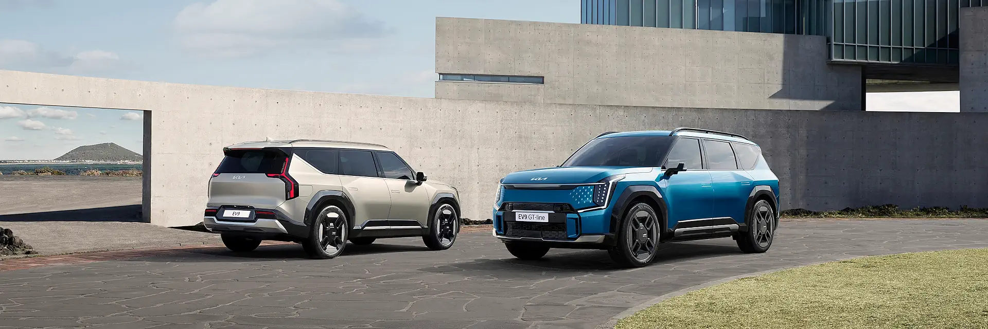 kia ev9 adw6 14 mobile Dwie Kie EV9 stoją na podjeździe przed betonową ścianą, beżowej widać tył i prawy bok, a niebieskiej przód i lewy bok