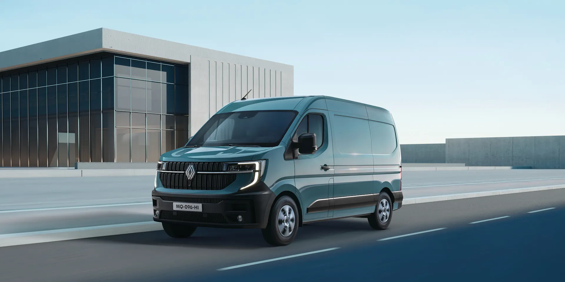 renault master adw1 02 Niebieski Renault Master jedzie na tle szarego nowoczesnego budynku, widać przód i lewy bok auta.