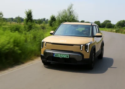 Żółta Kia EV2 jedzie drogą na tle krzaków, widać przód i lewy bok samochodu
