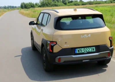 Żółta Kia EV2 jedzie drogą na tle krzaków, widać tył i lewy bok samochodu