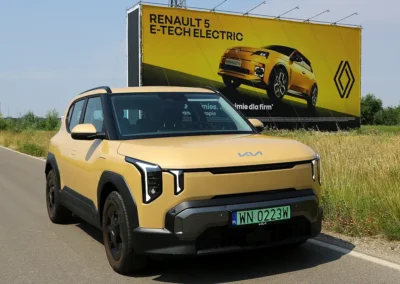Żółta Kia EV2 jedzie drogą na tle bilbordu renault, widać przód i prawy bok samochodu