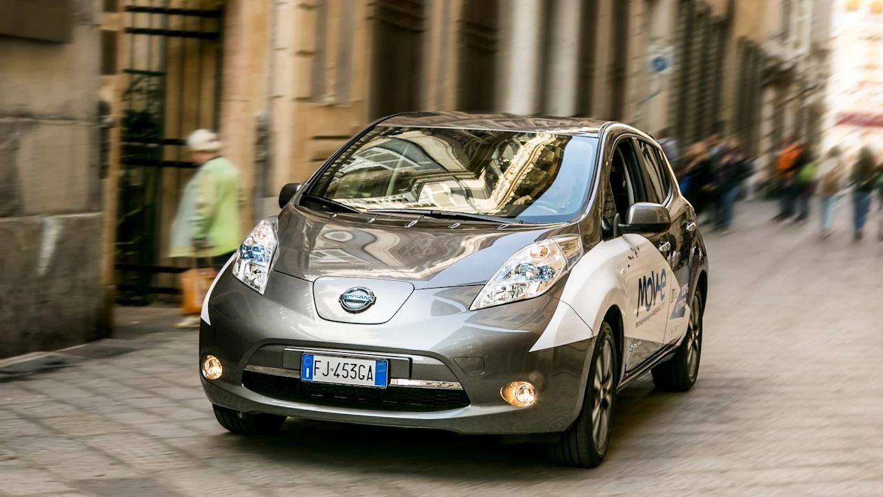 używane Nissan Leaf gen 1 4 Grafitowy Nissan Leaf jedzie przez miasto, widać przód i lewy bok auta