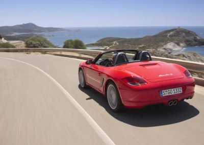 Czerwone Porsche Boxter 987 jedzie drogą na tle oceanu, widać tył i lewy bok auta
