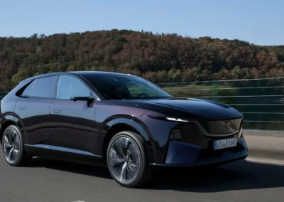 Fioletowa Mazda CX-6e jedzie asfaltową drogą – widać przód i prawy bok auta, w tle są las i niebo.