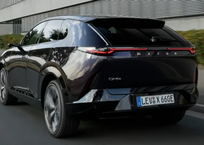 Fioletowa Mazda CX-6e jedzie asfaltową drogą przez miasto – widać tył i lewy bok auta, w tle jest fragment budynku.