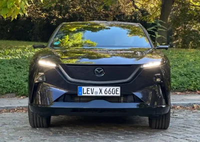 Fioletowa Mazda CX-6e stoi na brukowanej drodze – widać przód auta, w tle są drzewa.