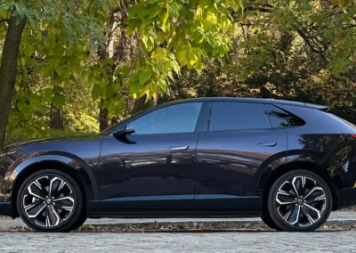 Fioletowa Mazda CX-6e stoi na brukowanej drodze – widać lewy bok auta, w tle są drzewa.