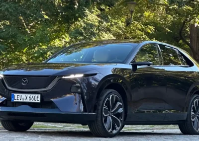 Fioletowa Mazda CX-6e stoi na brukowanej drodze – widać przód i lewy bok auta, w tle są drzewa.