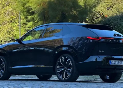 Fioletowa Mazda CX-6e stoi na brukowanej drodze – widać tył i lewy bok auta, w tle są drzewa.