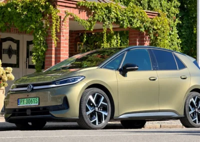 Volkswagen ID.3 Neo stoi na asfaltowej drodze – widać przód i lewy bok auta, w tle jest fragment budynku porośniętego bluszczem.