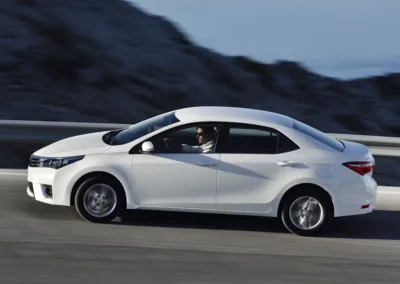 biała Toyota Corolla 11 generacji (e160) jadąca drogą wśród skał. Widok na lewy profil auta